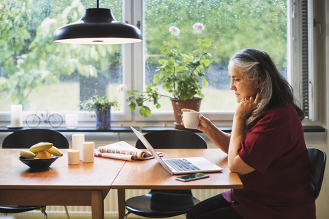 Seitenansicht einer älteren Frau, die einen Laptop benutzt, während sie am Tisch Kaffee trinkt, lizenzfreies Stockfoto