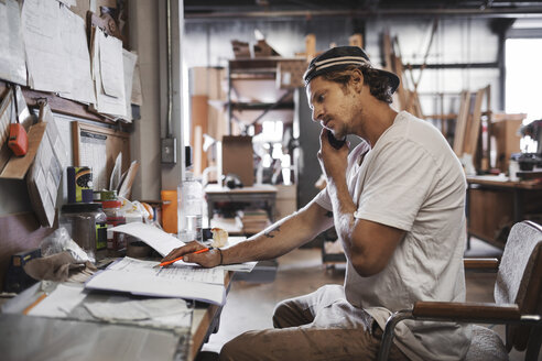 Seitenansicht eines Tischlers, der ein Mobiltelefon benutzt, während er ein Dokument in der Werkstatt liest - MASF03966