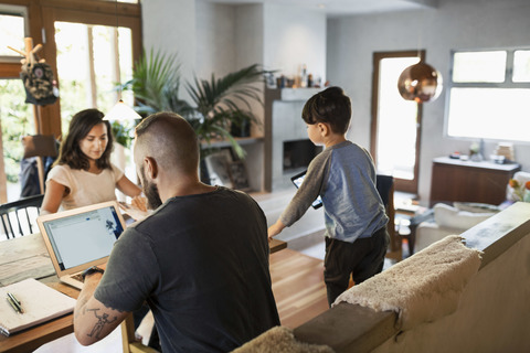Rückansicht eines Mannes, der an einem Laptop arbeitet, während die Familie am Esstisch im Haus sitzt, lizenzfreies Stockfoto