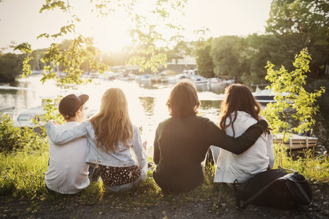 Rear view of teenagers sitting with arms around at lakeshore stock photo