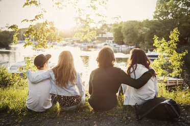 Rear view of teenagers sitting with arms around at lakeshore - MASF03991