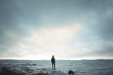 Rückansicht von Wonderlust Frau steht auf Felsen am Meer gegen bewölkten Himmel, lizenzfreies Stockfoto