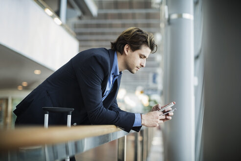 Seitenansicht eines Geschäftsmannes, der ein Mobiltelefon benutzt, während er sich am Geländer eines Flughafens abstützt - MASF04018
