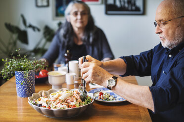 Senior man serving salad for himself while sitting with woman at table - MASF04033