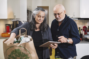 Senior couple with grocery bag using digital tablet in kitchen - MASF04038