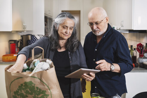 Älteres Paar mit Einkaufstasche und digitalem Tablet in der Küche - MASF04038