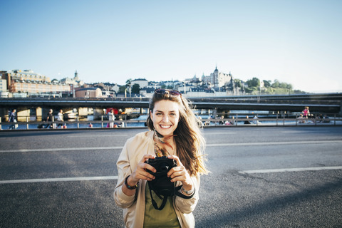 Porträt eines glücklichen Touristen, der auf einer Brücke gegen den klaren Himmel eine Kamera hält, lizenzfreies Stockfoto