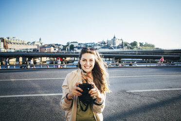 Porträt eines glücklichen Touristen, der auf einer Brücke gegen den klaren Himmel eine Kamera hält - MASF04086
