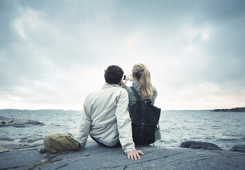 Rückansicht eines Paares, das ein Selfie macht, während es auf einem Felsen am Meer sitzt - MASF04095