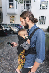 Mittlerer Erwachsener, der ein Mobiltelefon benutzt, während er ein Baby auf dem Bürgersteig trägt - MASF04195