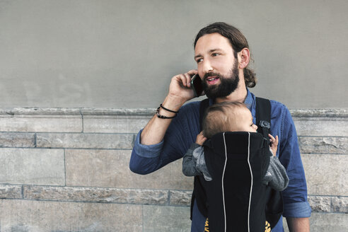 Mid adult man using mobile phone while carrying son in baby carrier against wall - MASF04197