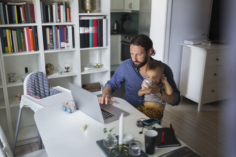 Mid adult father feeding baby boy while using laptop at home stock photo