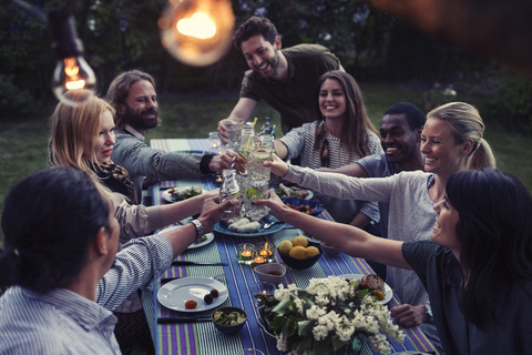 Fröhliche multiethnische Freunde stoßen mit Getränken am Esstisch im Hof an, lizenzfreies Stockfoto