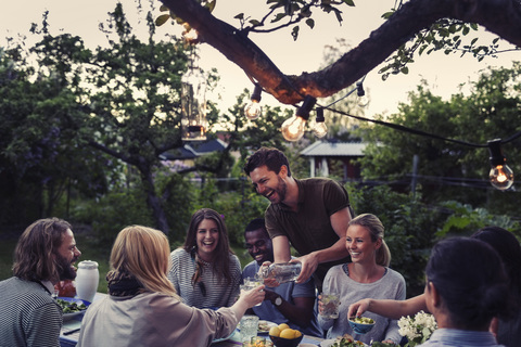 Glücklicher Mann, der einem Freund während einer Dinnerparty im Hof Wasser serviert, lizenzfreies Stockfoto