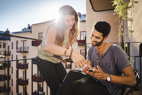 Glückliches Paar, das auf dem Balkon ein Smartphone benutzt und einen Reiseführer liest - MASF04258
