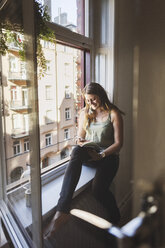 Happy woman using smart phone while holding guidebook on window sill at home - MASF04261