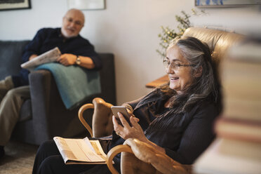 Happy senior woman using mobile phone while man sitting on sofa at home - MASF04274