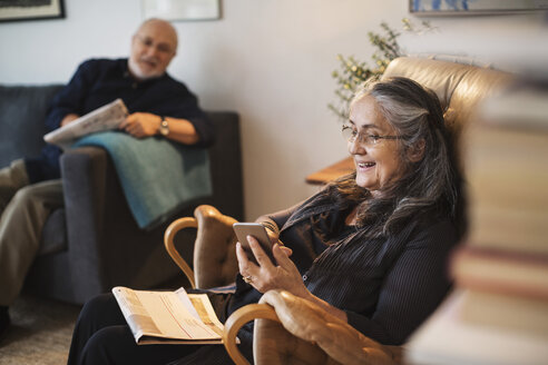 Glückliche ältere Frau, die ein Mobiltelefon benutzt, während der Mann zu Hause auf dem Sofa sitzt - MASF04274