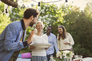 Glückliche junge Frau, die einen Teller hält, während sie ein Sommerfest mit Freunden im Garten genießt - MASF04314