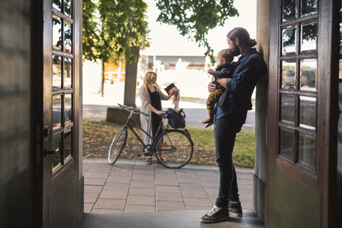 Father at door holding baby while looking at woman leaving for work stock photo