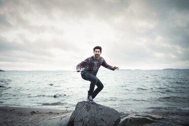 Full length portrait of happy man on rock by sea against cloudy sky - MASF04424