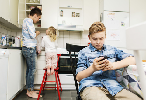 Junge benutzt Mobiltelefon, während Mutter und Schwester in der Küche arbeiten, lizenzfreies Stockfoto