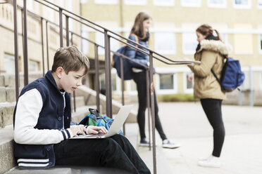 Junge, der einen Laptop auf einer Treppe benutzt, mit Schulmädchen im Hintergrund vor dem Schulgebäude - MASF04466