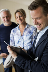 Smiling male real estate agent writing on document with couple standing in background - MASF04506