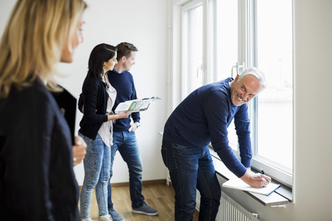 Smiling mature real estate agent looking at colleague white couple looking through window at home stock photo