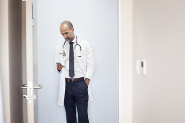 Arzt, der ein Smartphone benutzt, während er im Krankenhaus an der Wand steht, gesehen durch die Tür - CAVF38473