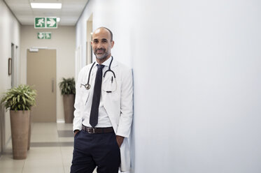 Portrait of smiling doctor with hands in pockets leaning on wall at hospital - CAVF38474