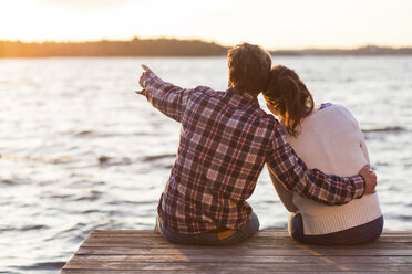 Rear view of man showing something to woman while sitting on pier against sea at sunset - MASF04649