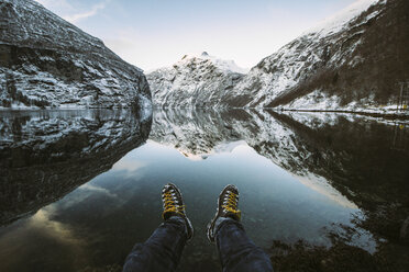 Low section of man sitting by at lakeshore by snowcapped mountains - CAVF38902