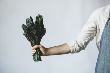 Midsection of man holding kale while standing against white wall at home - CAVF38964