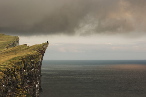 Entfernte Ansicht eines Mannes, der auf einer Klippe am Meer sitzt und die Aussicht betrachtet, lizenzfreies Stockfoto