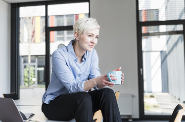 Lächelnde Frau mit einer Tasse Kaffee auf einem Tisch im Büro sitzend - UUF13343