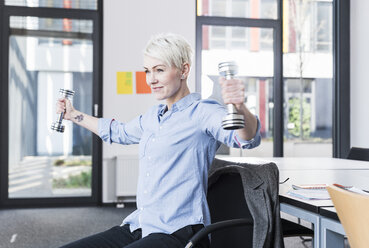 Smiling woman exercising with dumbbells in office - UUF13362