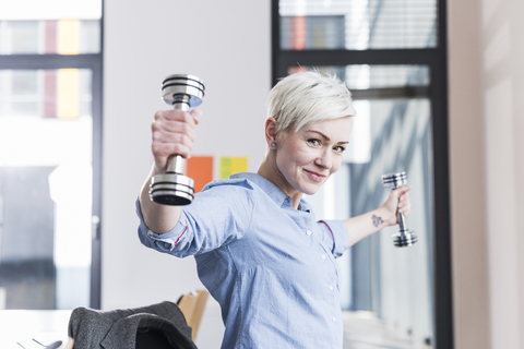 Porträt einer lächelnden Frau, die im Büro mit Hanteln trainiert, lizenzfreies Stockfoto