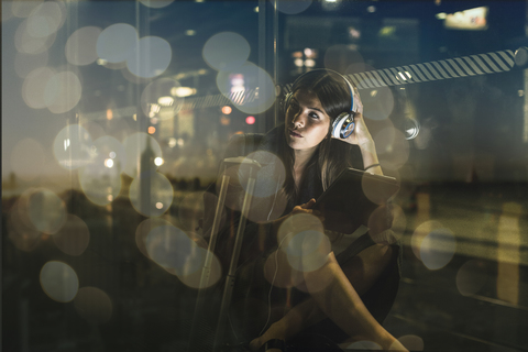 Young woman with tablet and headphones sitting outdoors at night stock photo