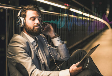 Young man with tablet and headphones sitting on bridge at night - UUF13407