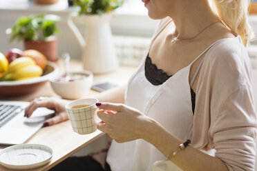 Frau mit Kaffee in der Hand, die einen Laptop benutzt, während sie am Tisch sitzt - CAVF39185