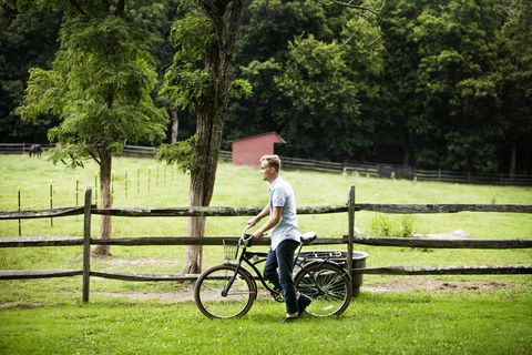 Mann mit Fahrrad geht am Zaun eines Bauernhofs vorbei, lizenzfreies Stockfoto