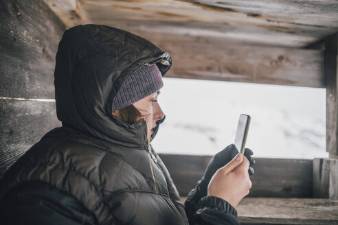 Österreich, Kitzbühel, junge Frau in Hochsitz schaut auf Handy im Winter - GUSF00632