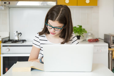 Mädchen mit Laptop und Blick auf ein Buch in der Küche - LVF06867