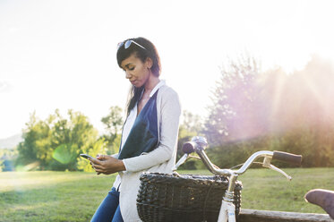 Smiling woman using phone while sitting on bench at park - CAVF39438