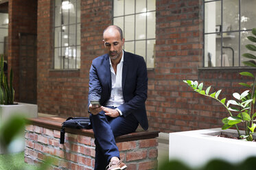 Businessman using smart phone while sitting on seat against office building - CAVF39444