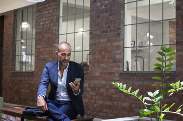 Geschäftsmann, der ein Smartphone benutzt, während er auf dem Sitz vor dem Büro sitzt - CAVF39445