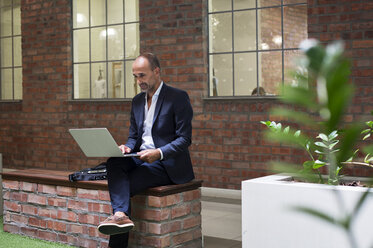 Geschäftsmann, der einen Laptop benutzt, während er auf einem Stuhl vor einem Bürogebäude sitzt - CAVF39446