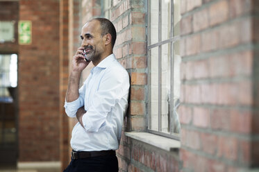 Side view of businessman talking on smart phone while standing against brick wall - CAVF39450