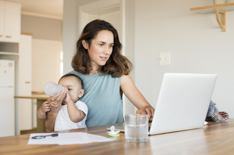 Frau füttert ihren Sohn, während sie einen Laptop am Tisch benutzt, lizenzfreies Stockfoto